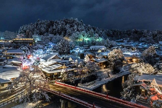 An elevated night view of snow-covered Takayama old town, with the red Nakabashi bridge lit up over the Miyagawa River