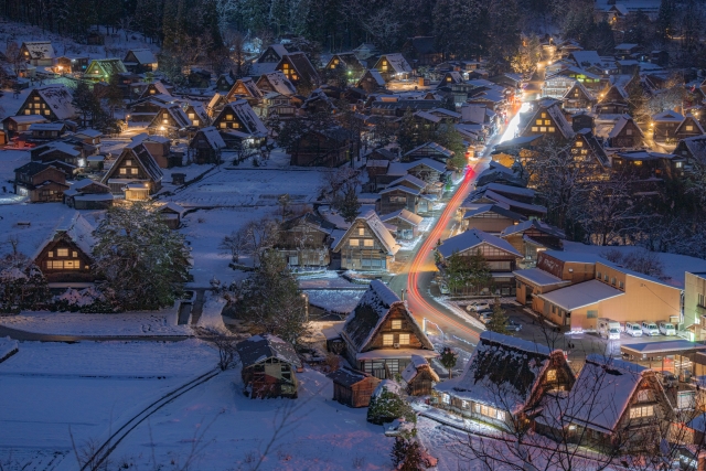 An elevated night view of snow-covered Shirakawa-go, with lit gassho-zukuri farmhouses and light trails from a passing car along the village road